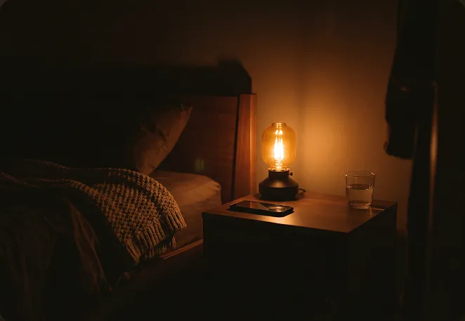 A bedside lamp lit beside a phone playing at low volume